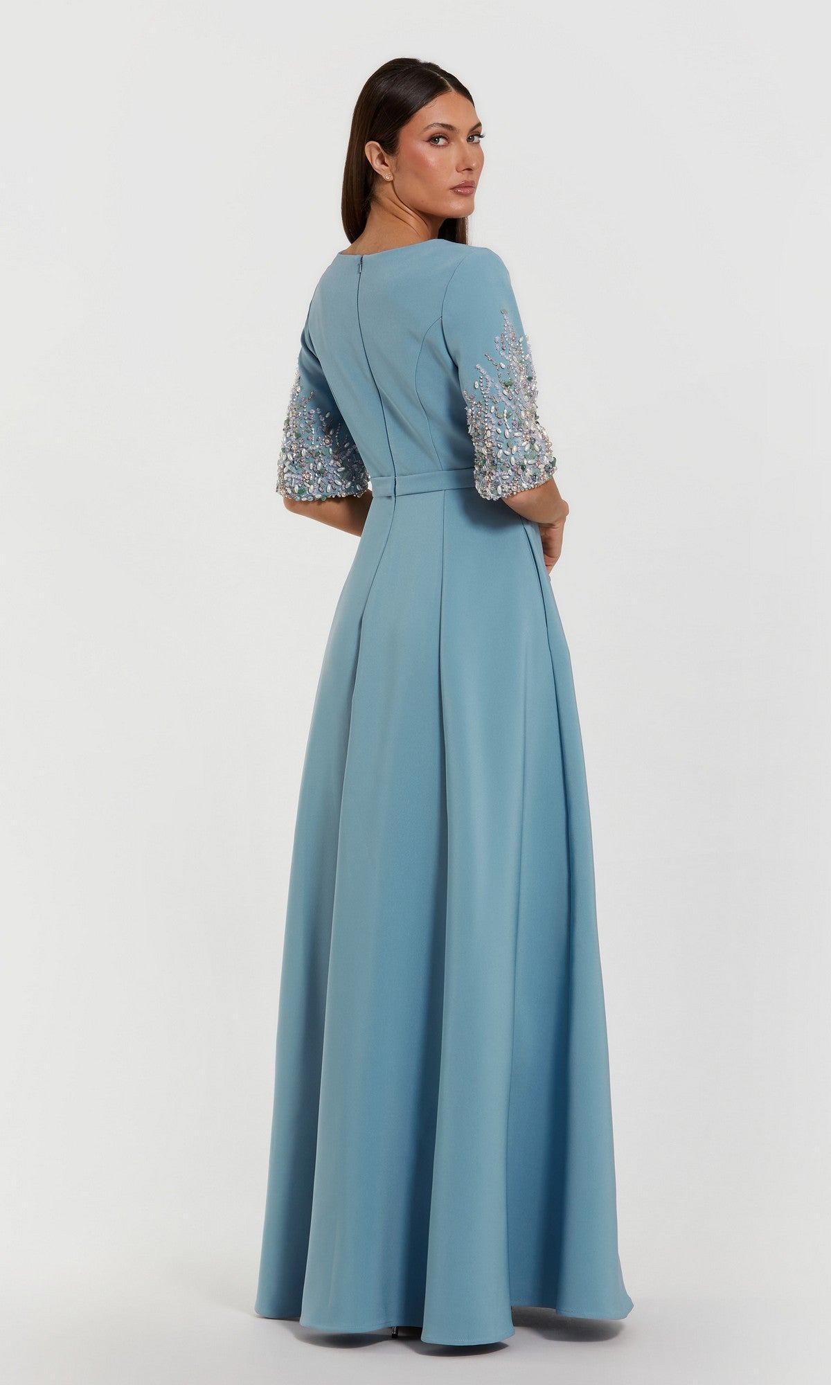 A woman in the Mac Duggal 2322 long formal dress, a floor-length light blue crepe gown with beaded floral embellishments on the sleeves, stands looking over her shoulder.